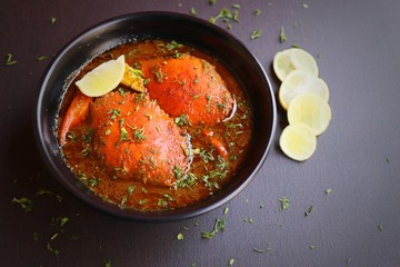 Indian Cuisine, Spicy Crabs Curry in red gravy, garnished with fresh coriander and lemon wedges or lemon slices on dark moody background