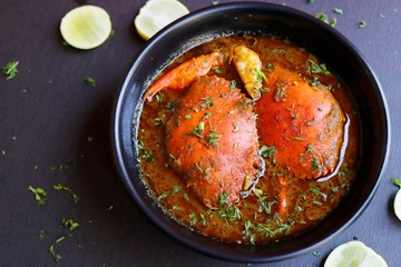 Indian Cuisine, Spicy Crabs Curry in red gravy, garnished with fresh coriander and lemon wedges or lemon slices on dark moody background