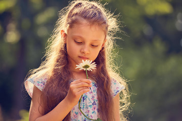 Beautiful kid girl looking on chamomile flower on nature summer green background. Closeup portrait