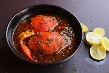 Indian Cuisine, Spicy Crabs Curry in red gravy, garnished with fresh coriander and lemon wedges or lemon slices on dark moody background
