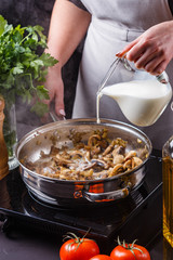 young woman in a gray apron adds milk to mushroom sauce