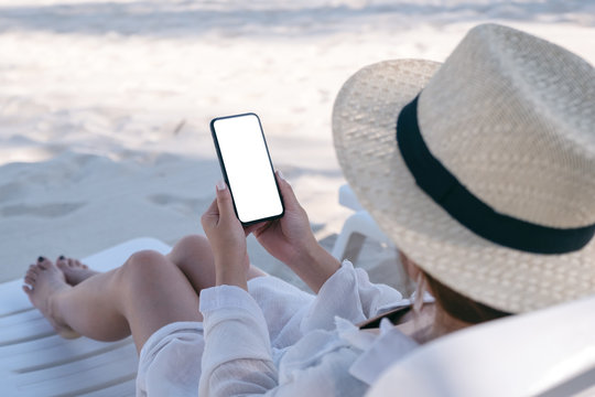 A woman holding white mobile phone with blank desktop screen while laying down on beach chair on the beach