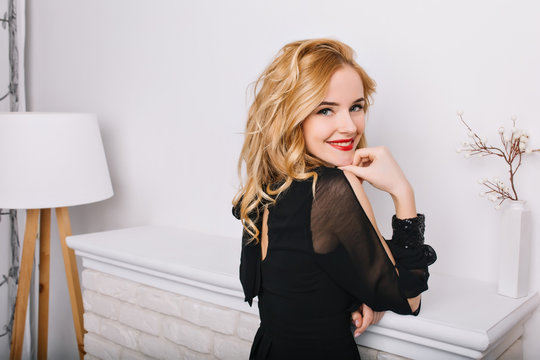 Side view closeup portrait of girl with blonde wavy hair in white modern room, studio with floor lamp and fireplace. Lady smiling to camera, posing. Wearing stylish black dress.