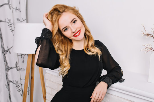 Attractive young woman with beautiful smile, sensually touching her wavy blonde hair in white modern room. Wearing fashineble black dress, red lipstick, light makeup.
