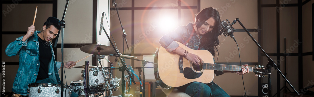 Wall mural panoramic shot of attractive woman playing guitar while mixed race musician playing drums in recording studio