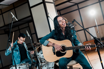 Fototapeta na wymiar selective focus of beautiful inspired musician playing guitar and singing while mixed race man playing drums in recording studio