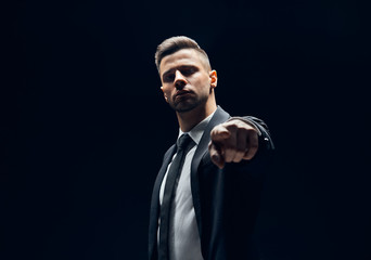Fototapeta na wymiar Serious handsome man in black suit pointing his finger to you and camera isolated on dark background