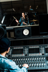 Fototapeta na wymiar selective focus of mixed race sound producer working at mixing console while pretty woman singing in recording studio