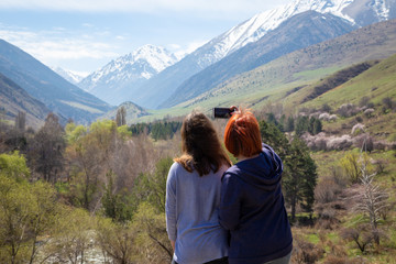 Two girls happily photographed against the backdrop of the mountains. Travelers take a selfie on the background of a beautiful mountain gorge. High snowy mountains and green trees in front of them.