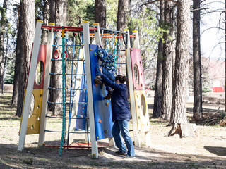 Mother helps her son climb the wall in the playground outside on a sunny day. Trees in the background.