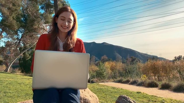 A happy, attractive, young, caucasian woman on her laptop at the park, sitting on a rock - she smiles and lets out a little laugh. Beautiful scenery. Slow motion - No logo - Steadicam - Close up.