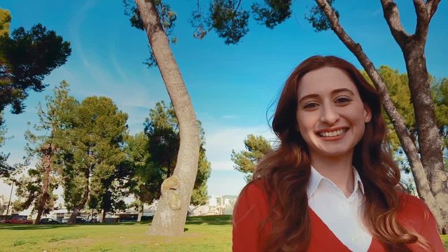 A happy, attractive, young, caucasian woman at the park, turns her head to the camera and lets out a big smile. Slow motion - Steadicam - Close up.