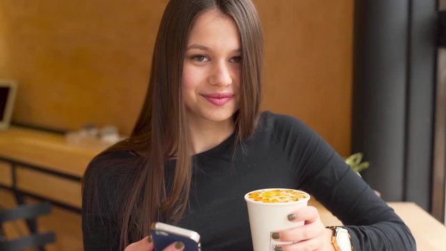 Young Woman Holds Smartphone And Drink And Winks At Camera In Coffee Shop