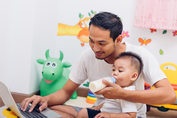 Father acting Mom feeding milk his son baby 1 year old while working on laptop computer