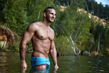 Brutal pumped guy in sexy swimming trunks stands smiling in the lake