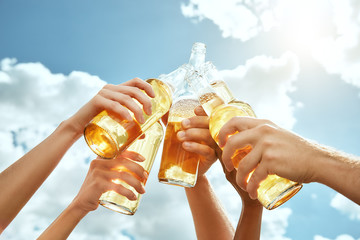 Beer time! Cropped image of hands are clinking bottles with beer against summer sky background....