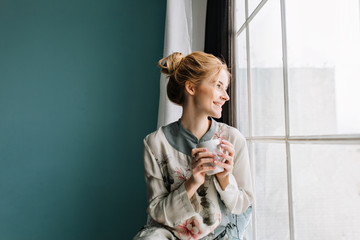 Portrait of young woman with blonde hair drinking coffee or  tea next to big window, smiling, enjoying happy morning at home. Turquoise wall on background. Wearing silk pajamas in flowers.