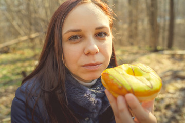 Young woman eating doughnut in woods Casual young brunette in jacket sitting on log in woods and eating delicious glazed doughnut