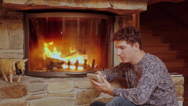 Man Sits by a Cozy Fire and Writes on His Notepad.