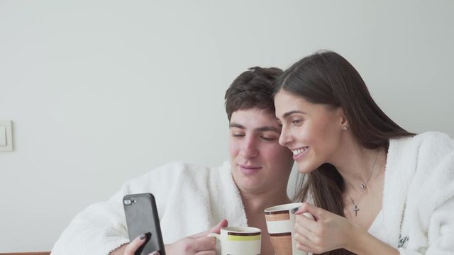 Happy couple in bathrobes drinking coffee and taking photos of themselves