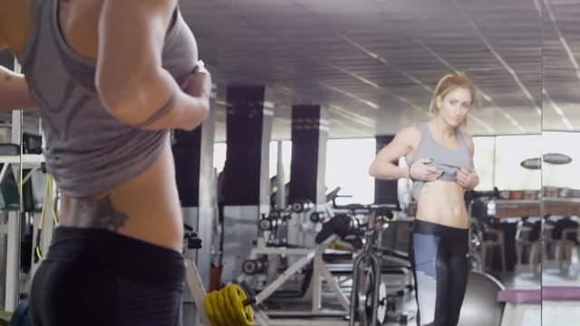 Slow motion clip of a fit woman at the gym as she holds up her shirt looking at her stomach in the mirror