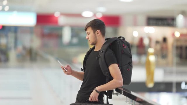 A handsome bunet boy has just come back from a trip and stops at the mall to find Wi-Fi in order to inform his parents and girlfriend that he is safe and sound. The background is blurred.