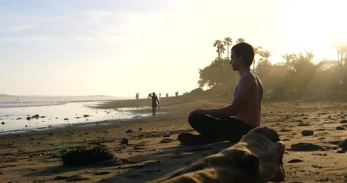 A handsome man deep breathing and meditating at sunset on a picturesque California beach with palm trees in silhouette.