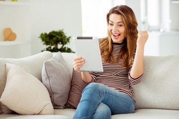 Portrait of her she nice-looking lovely attractive cheerful cheery ecstatic wavy-haired girl sitting on divan reading digital book yes luck in light white interior room