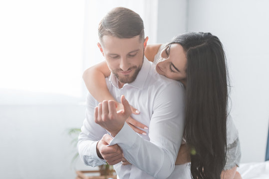 cheerful girlfriend hugging bearded boyfriend in white shirt at home