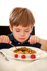 Hungry child sitting in chair at table in kitchen and eating with spoon of cooked peas with tomato. Kids meal and healthy diet food concept. Happy and cute little boy enjoying in good lunch at home.