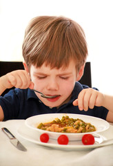 Hungry child sitting in chair at table in kitchen and eating with spoon of cooked peas with tomato. Kids meal and healthy diet food concept. Happy and cute little boy enjoying in good lunch at home.