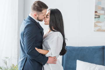 happy bearded man in suit kissing nose of attractive girlfriend in white shirt