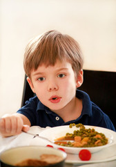 Hungry child sitting in chair at table in kitchen and eating with spoon of cooked peas with tomato. Kids meal and healthy diet food concept. Happy and cute little boy enjoying in good lunch at home.