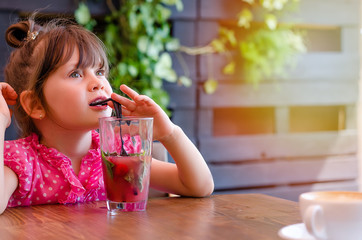 Adorable little girl drinking lemonade with raspberry and basil at table in cosy outdoor cafe....