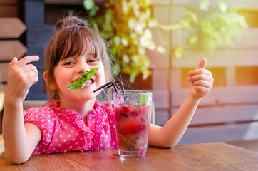 Adorable little girl drinking lemonade with raspberry and basil at table in cosy outdoor cafe. Happy and healthy childhood concept. Copy space - obrazy, fototapety, plakaty