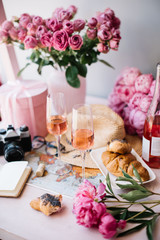 Obraz na płótnie Canvas old vintage film camera, paper notebook, world map, hat, croissants, pink peonies, roses in a vase, velvet pink gift box and a bottle of rose wine with two glasses on the pink wooden table 