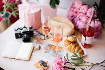 old vintage film camera, paper notebook, world map, hat, croissants, pink peonies, roses in a vase, velvet pink gift box and a bottle of rose wine with two glasses on the pink wooden table 