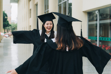 Young beautiful female graduate hugging best friend at graduation ceremony. Pretty asian college...