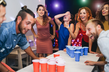 Happy friends playing beer pong in a cocktail bar - Young millennial people having fun doing party...