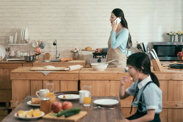 ignore family gadget addiction. busy asian wife mother holding wooden plate with croissant walking in modern kitchen talking with husband on cellphone in morning. little girl eating having breakfast