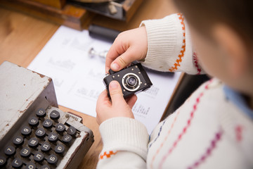 Old vintage spy camera in the hands of a little boy on the background of an old typewriter or computer. The boy understands the mechanism of the camera. Early development.