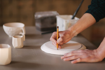 Fototapeta na wymiar Potter making ornament on ceramic plate. Professional male potter draw a pencil on ceramic product