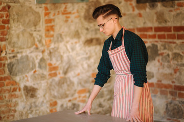 Young male artist in aprong and glasses stand in front of brick wall