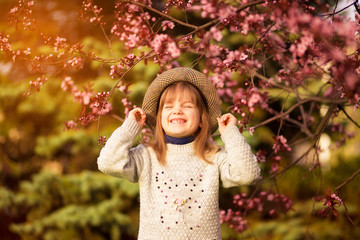 Spring portrait, adorable little girl in hat walk in blossom tree garden on sunset