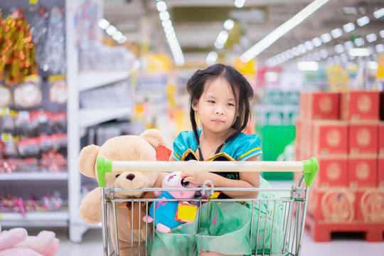 little asian girl in princess costume sitting with dolls in the cart between shopping