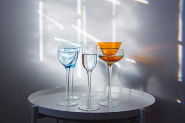 Group of tall long-stemmed cocktail glasses in different shapes and colours filled with clear liquid on round white table with geometric patterns made by rays of sunlight against grey background