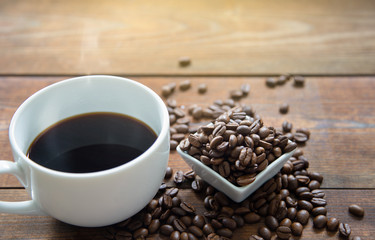 A white cup of hot coffee with coffee beans in white small ceramic dish on wooden table and morning sun light.