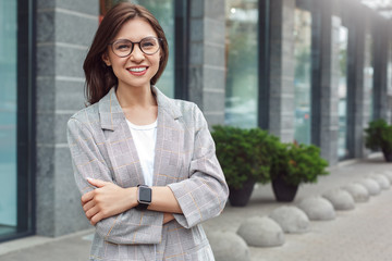 Business casual. Young lady in eyeglasses standing on the city street crossed arms smiling happy