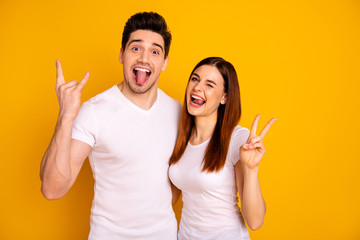 Close up photo beautiful amazing she her he him his guy lady pair hold hands arms  v-sign metal concert horns tongue out mouth wink eye carefree wear casual white t-shirts isolated yellow background