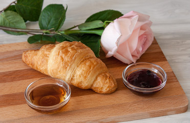One fresh crispy croissant flower pink roses, cups with PSAS maple and blueberry jam on a cutting wooden board on a light beige desk. 